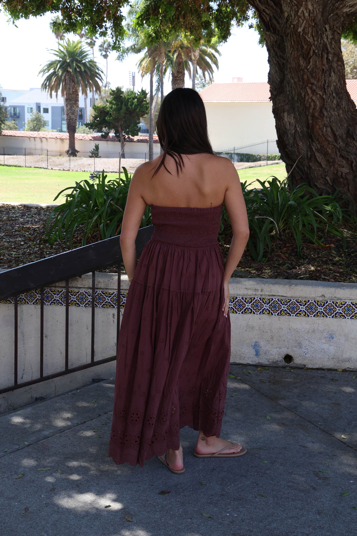 A woman stands outdoors on a paved walkway, viewed from behind, wearing a strapless maroon maxi dress with a gathered waist and tiered skirt. Trees, grass, and a low wall with decorative tile are visible in the background.