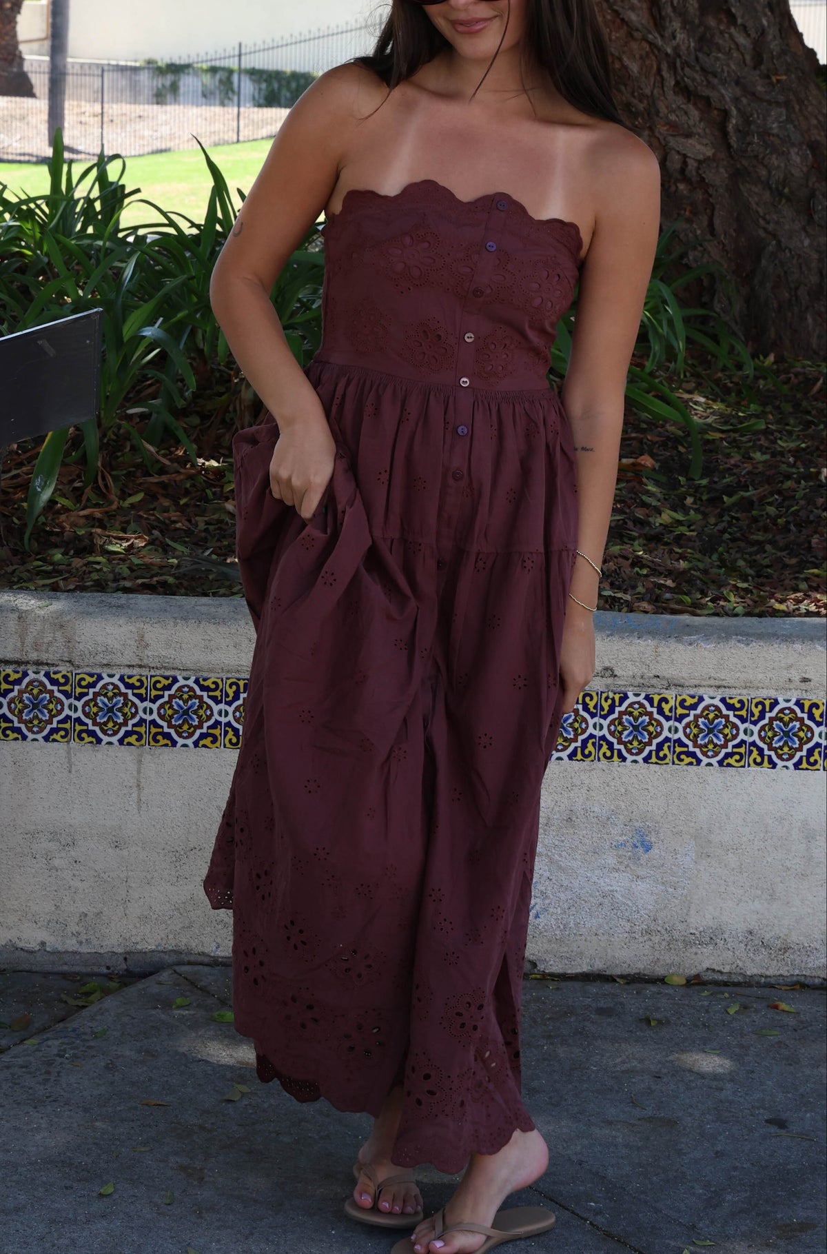 A woman stands outdoors on a paved walkway, viewed from behind, wearing a strapless maroon maxi dress with a gathered waist and tiered skirt. Trees, grass, and a low wall with decorative tile are visible in the background.