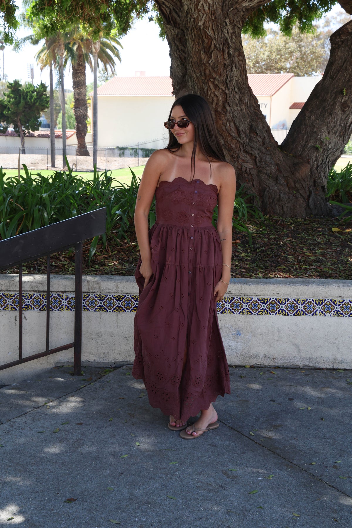 A woman stands outdoors on a paved walkway, viewed from behind, wearing a strapless maroon maxi dress with a gathered waist and tiered skirt. Trees, grass, and a low wall with decorative tile are visible in the background.