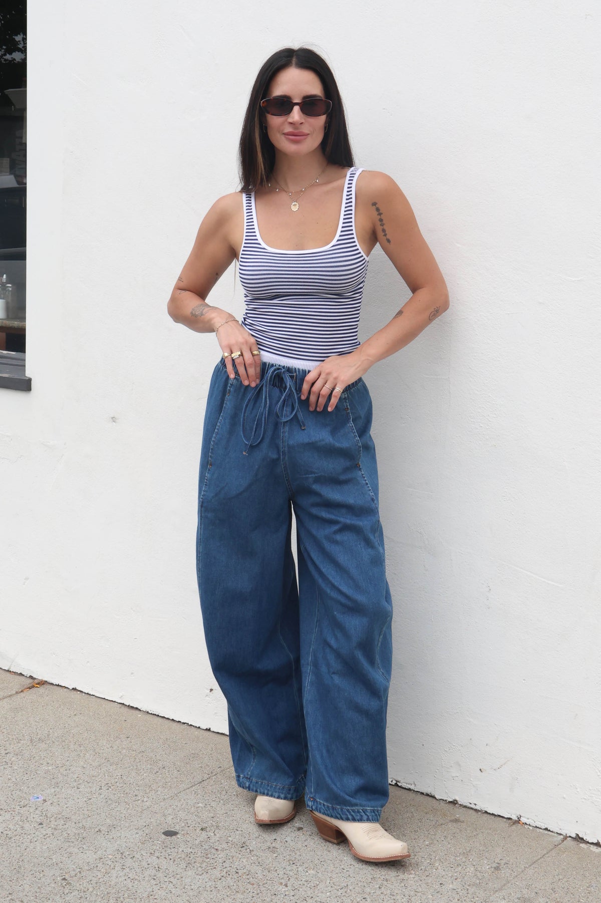 Woman wearing a striped tank top and blue wide-leg pants against a white wall.