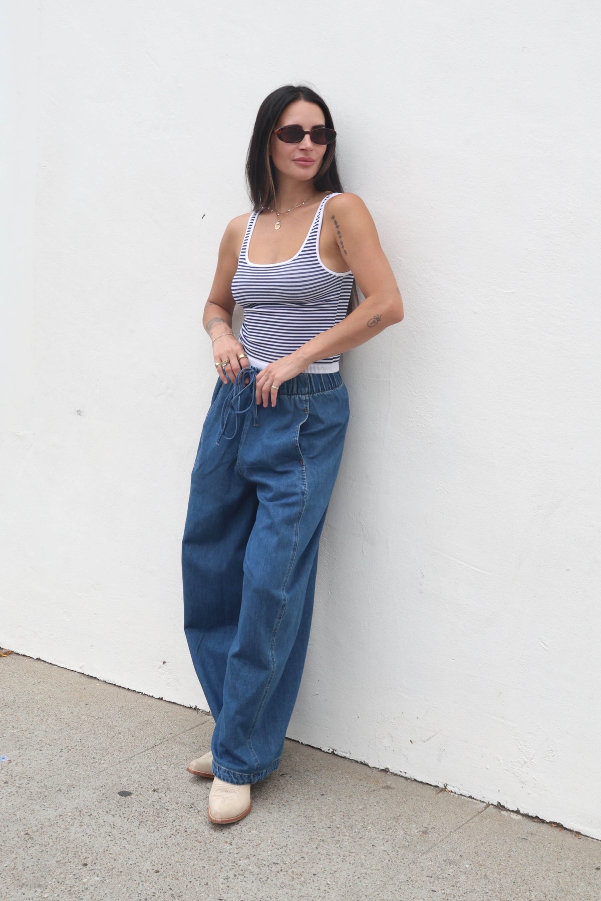 Woman wearing a striped tank top and blue jeans against a white wall.