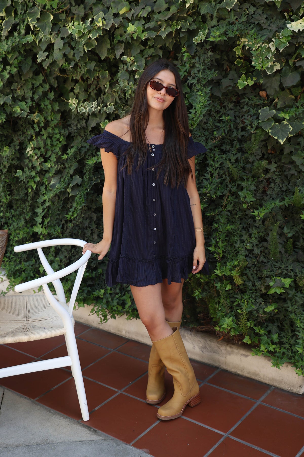 A woman wearing a navy blue off-the-shoulder ruffle dress and tan cowboy boots stands in front of a green ivy wall, holding onto a white chair.