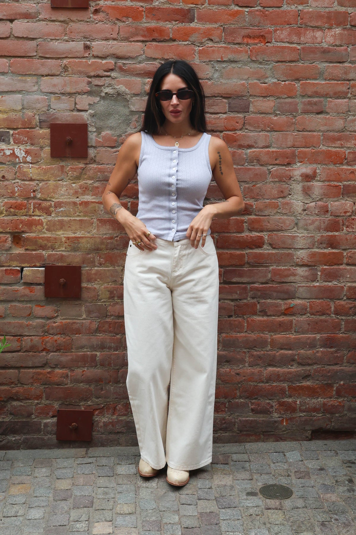 Woman wearing a light blue sleeveless top and white pants standing against a brick wall.