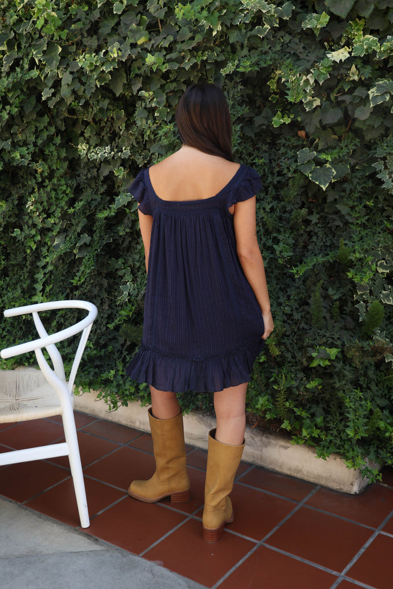 A woman stands with her back to the camera wearing a navy blue off-the-shoulder pleated mini dress and tan knee-high boots, posing in front of a green ivy-covered wall.