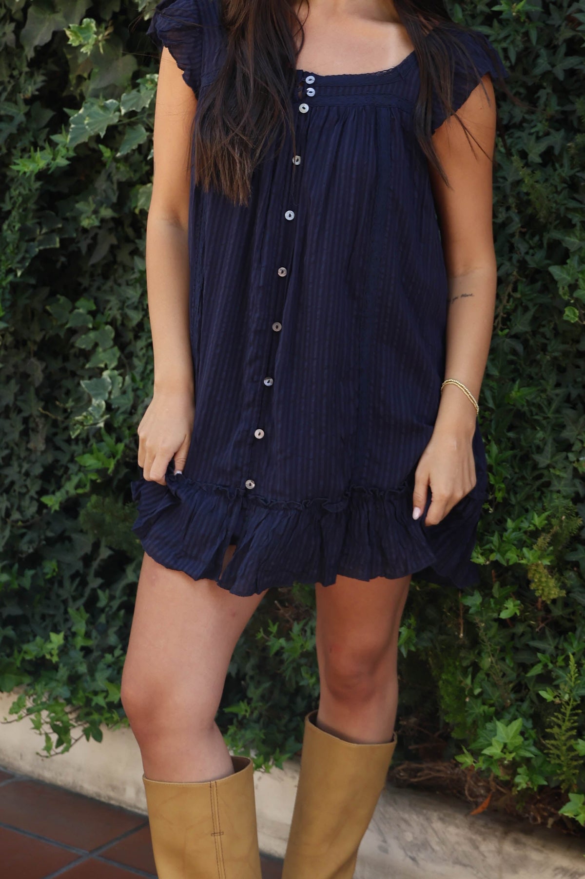 A woman wearing a navy blue short-sleeve button-up dress and tan knee-high boots stands outdoors in front of a green ivy wall, next to a white chair.