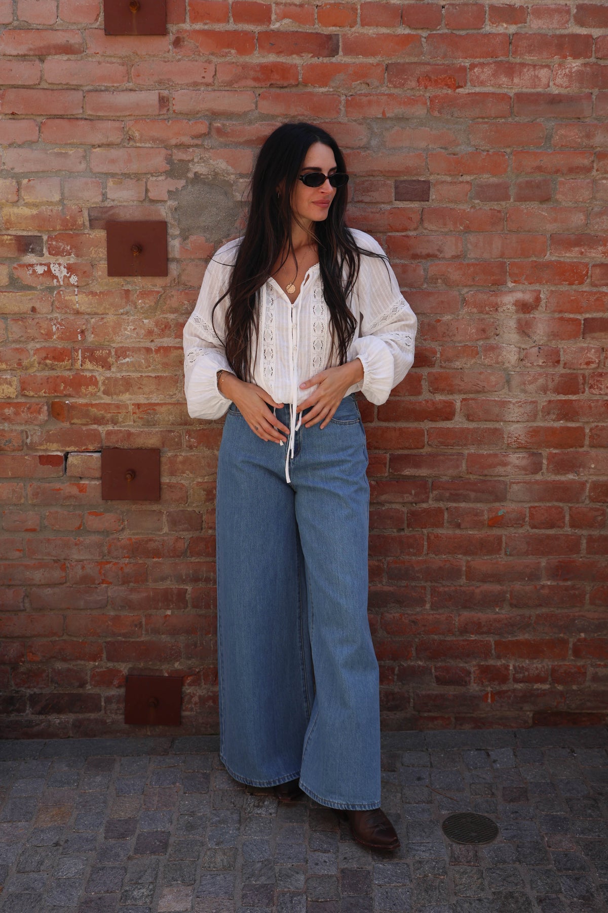 Woman with a white lace long sleeve detailed blouse against a brick wall