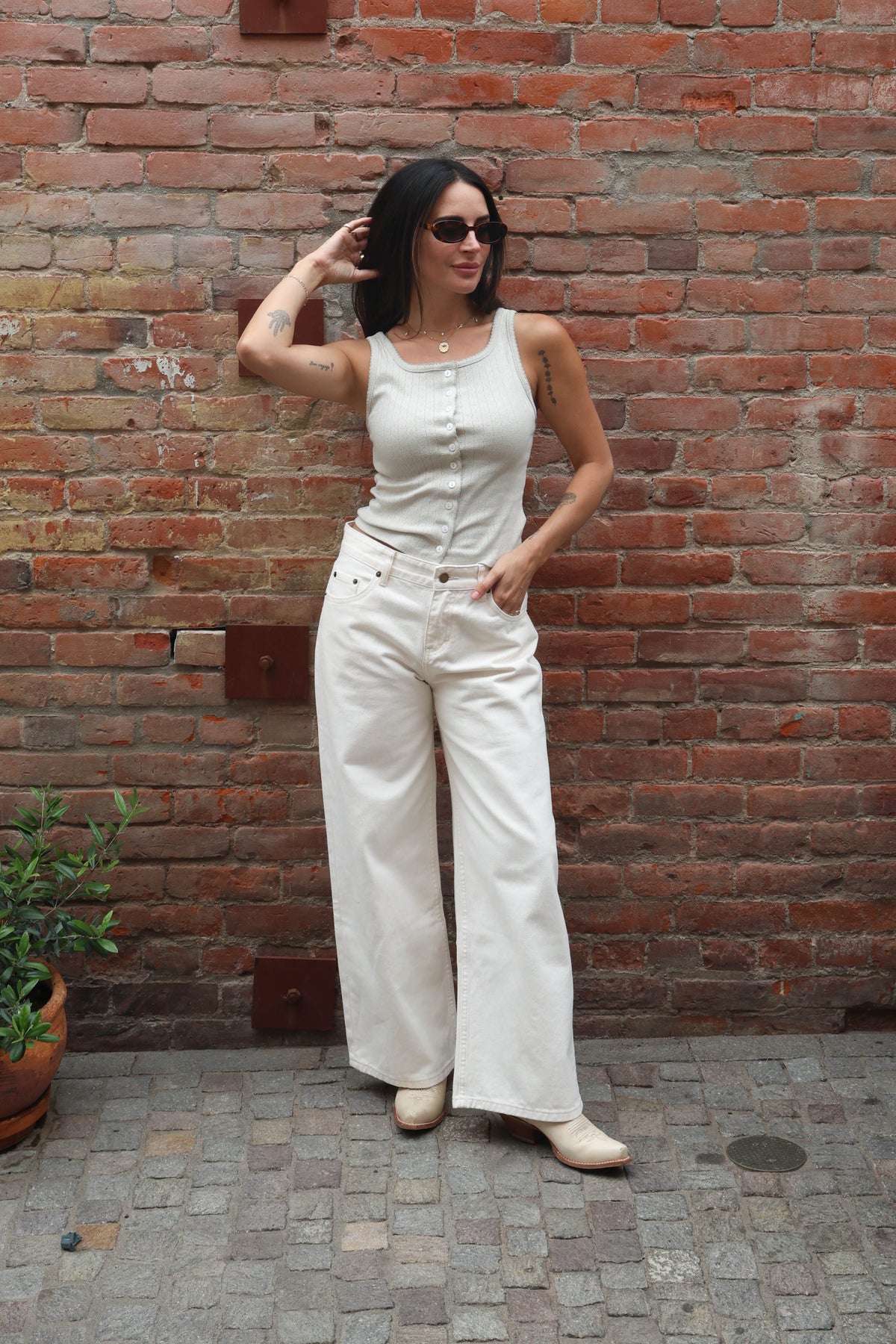 Woman in a white sleeveless top and wide-leg pants standing against a brick wall.