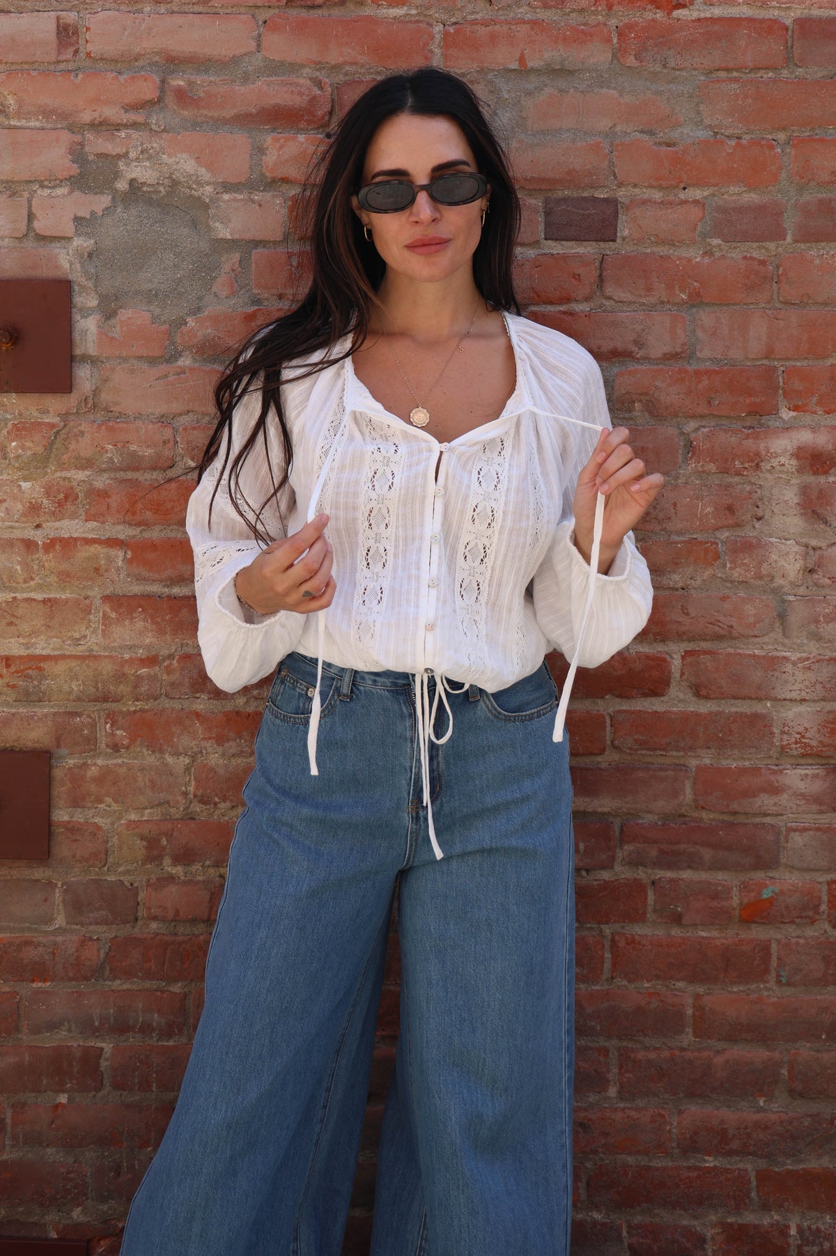 Woman with a white lace long sleeve detailed blouse against a brick wall