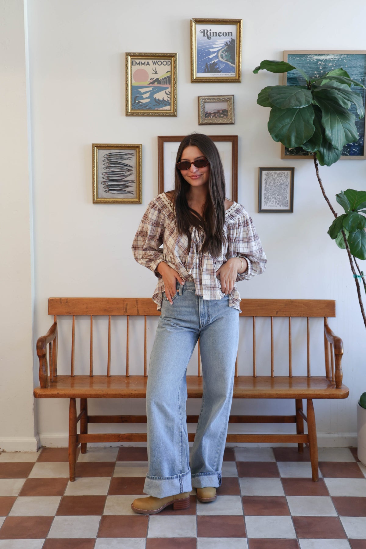 A woman wearing a cream-colored plaid blouse with brown and red lines, featuring a lace-trimmed scoop neckline and ruffled details, paired with light-wash blue jeans.
