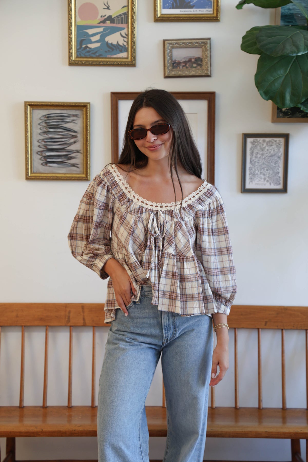 A woman wearing a cream-colored plaid blouse with brown and red lines, featuring a lace-trimmed scoop neckline and ruffled details, paired with light-wash blue jeans.