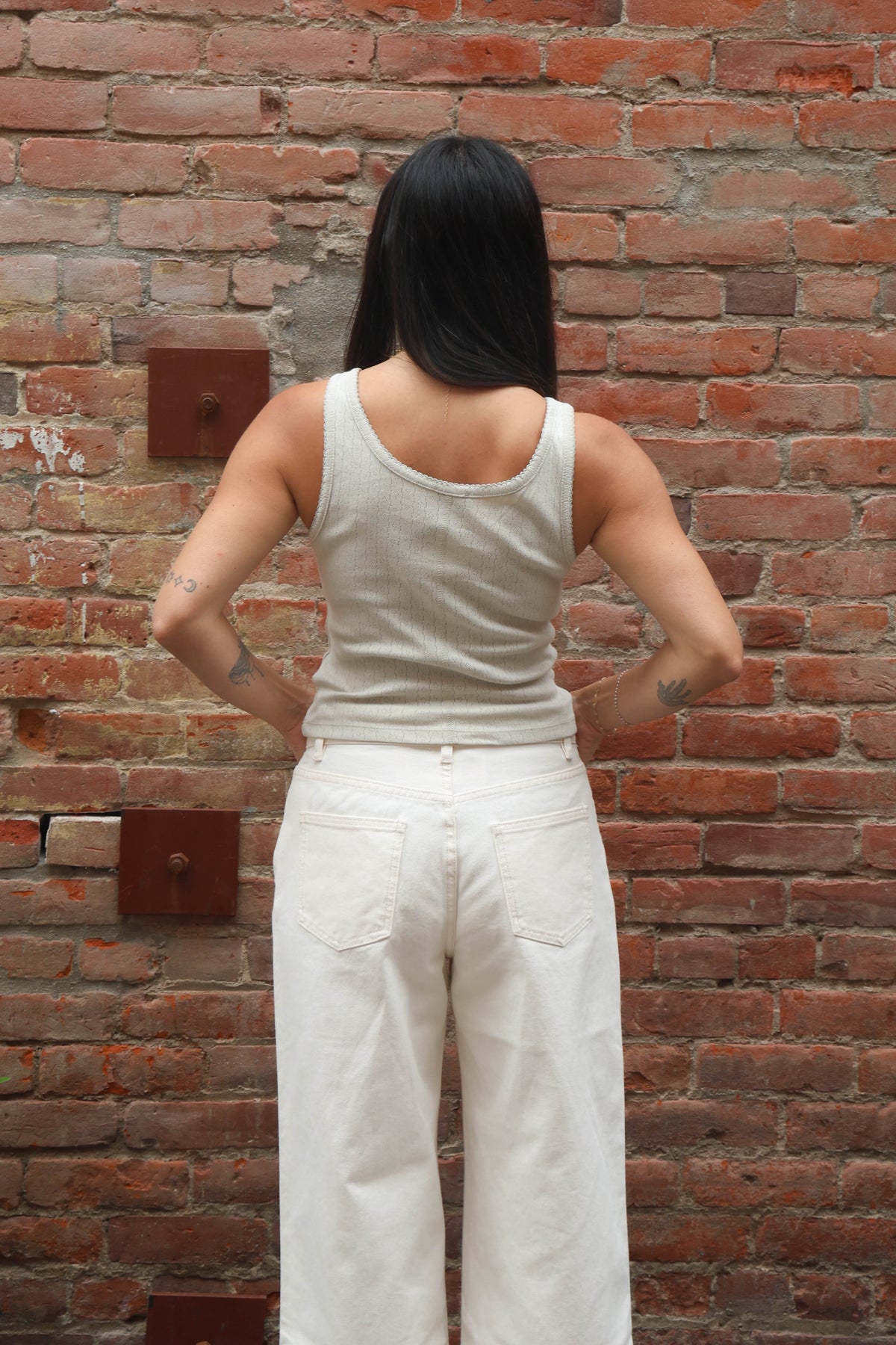 Person wearing a beige tank top and white pants standing against a brick wall.