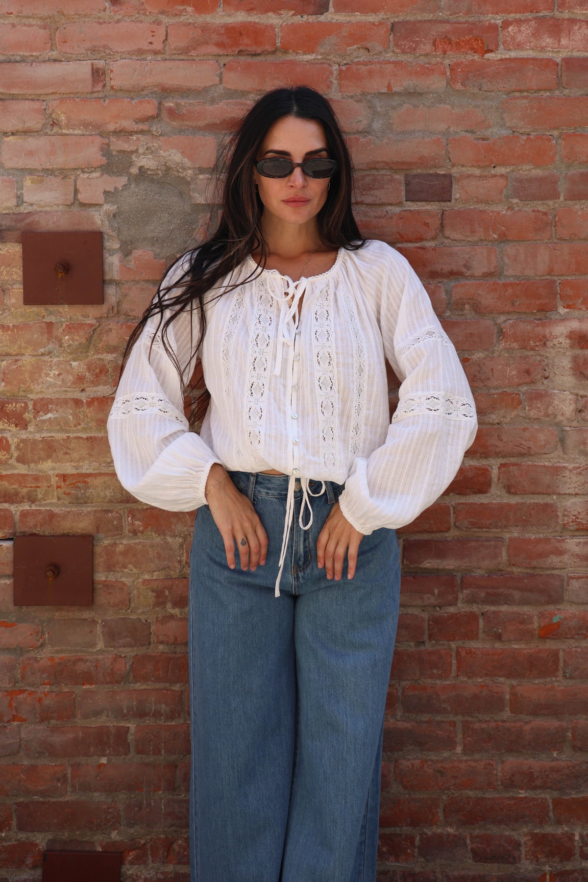 Woman with a white lace long sleeve detailed blouse against a brick wall
