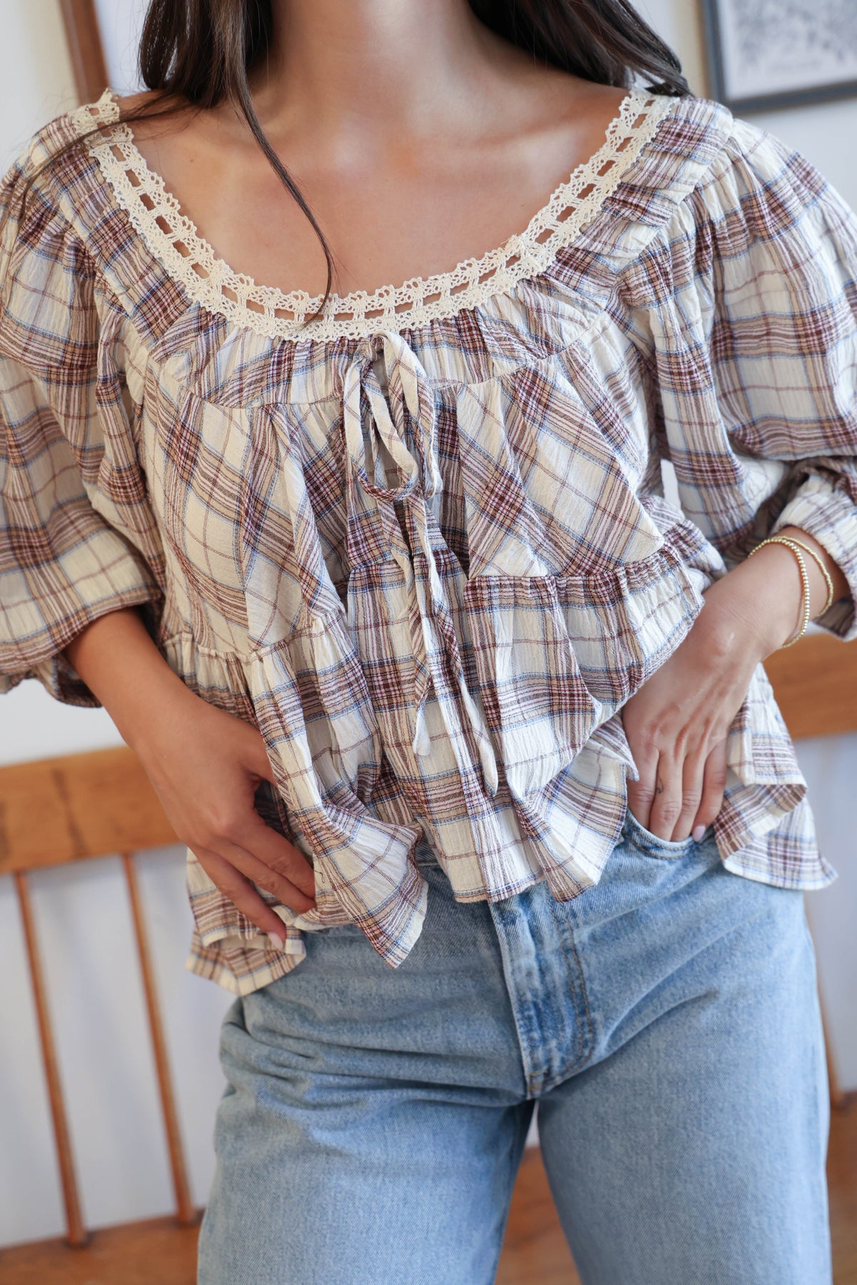 A woman wearing a cream-colored plaid blouse with brown and red lines, featuring a lace-trimmed scoop neckline and ruffled details, paired with light-wash blue jeans.