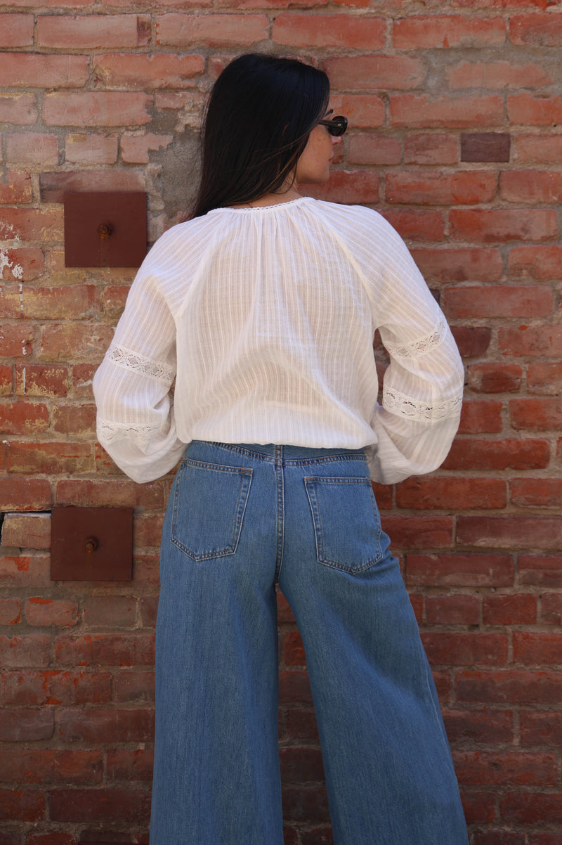 Woman with a white lace long sleeve detailed blouse against a brick wall