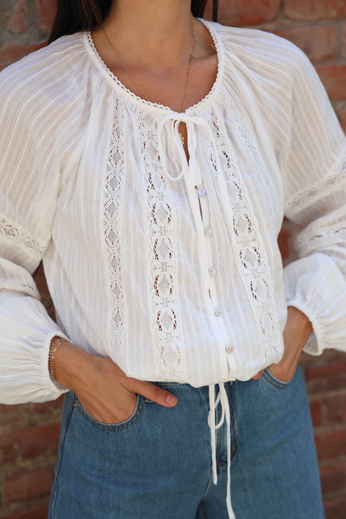 Woman with a white lace long sleeve detailed blouse against a brick wall