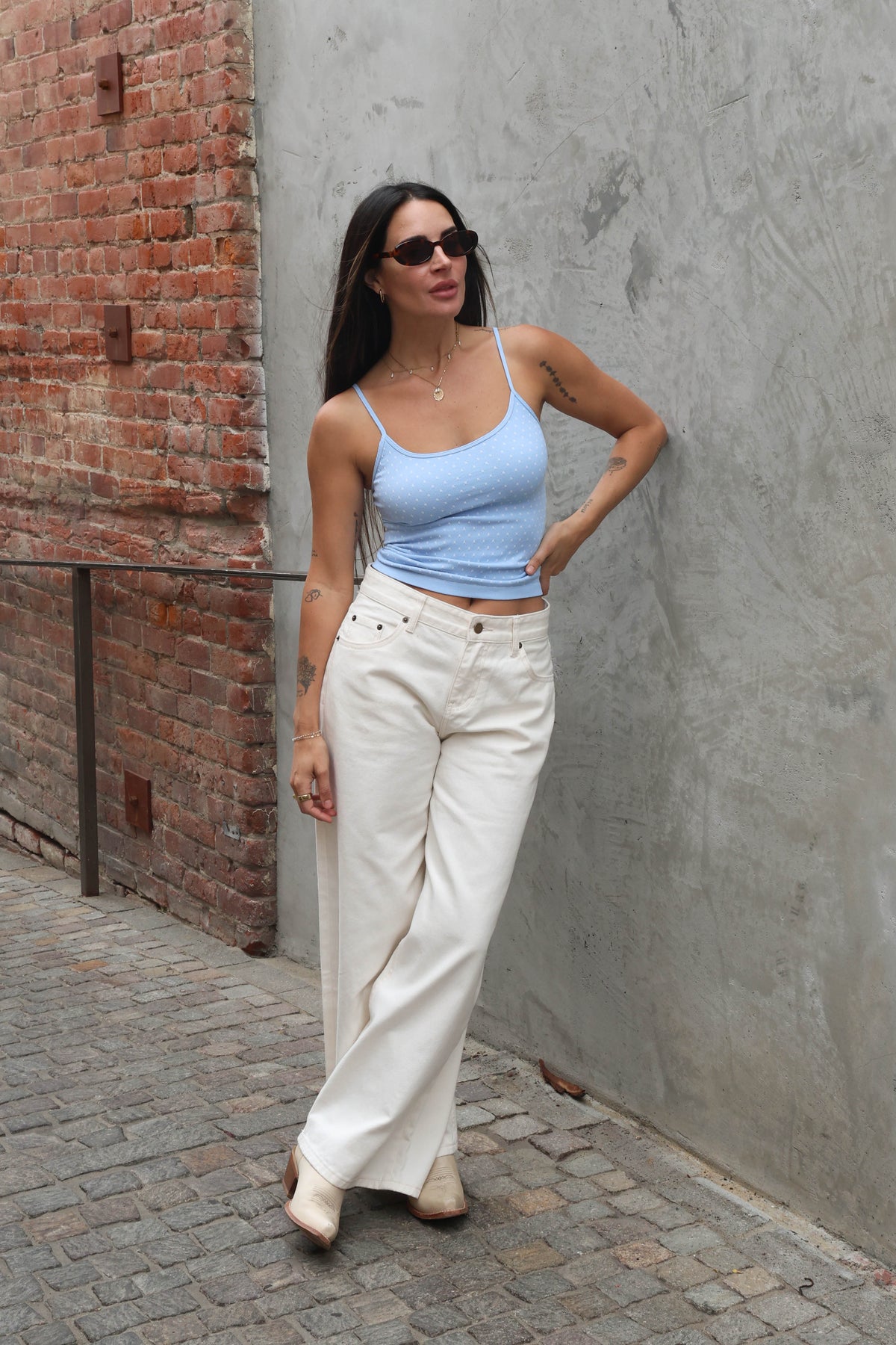 Woman wearing a light blue tank top and white pants standing against a gray wall.