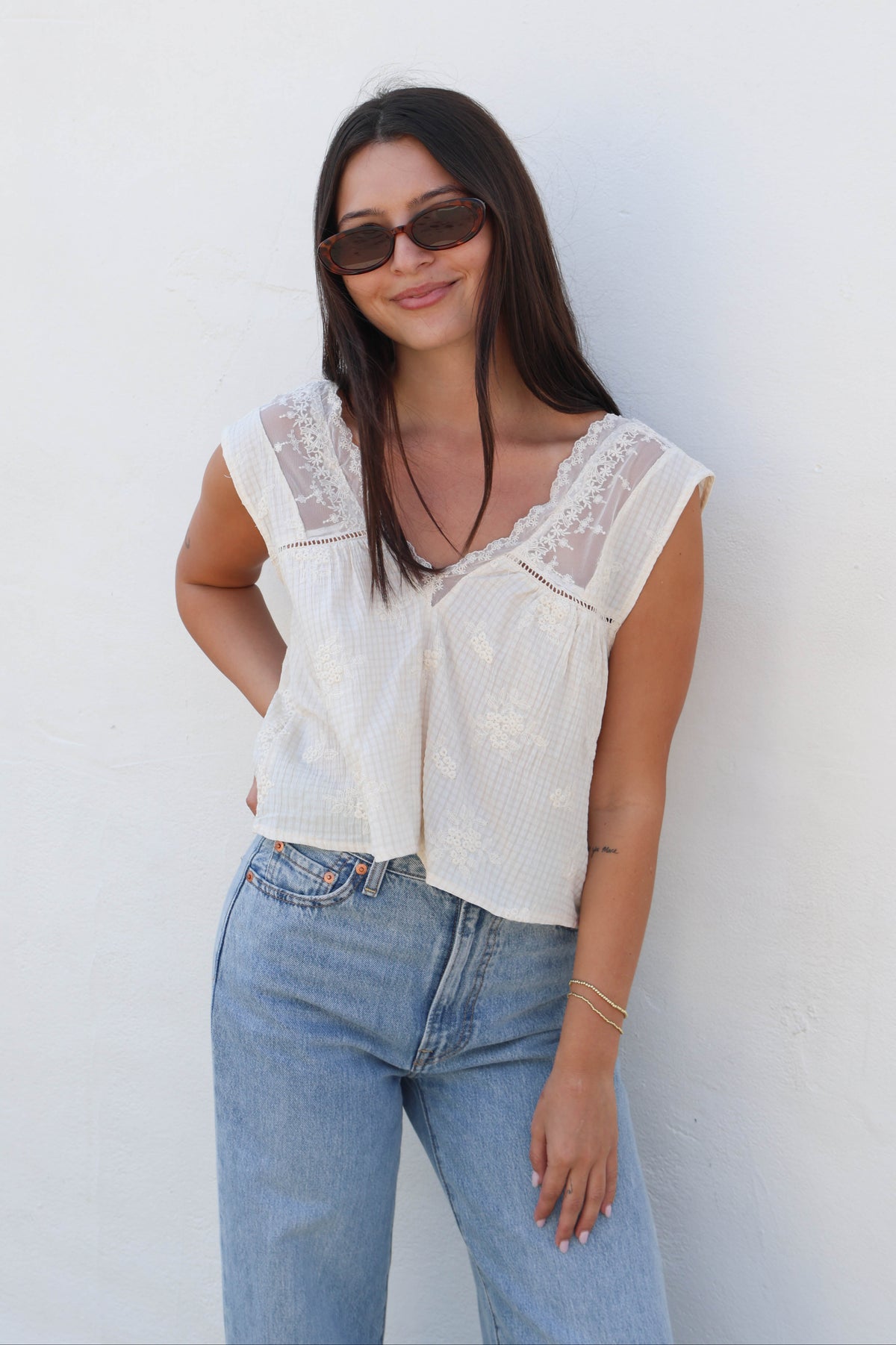 Woman wearing a white tank with lace detail with jeans against a white wall.