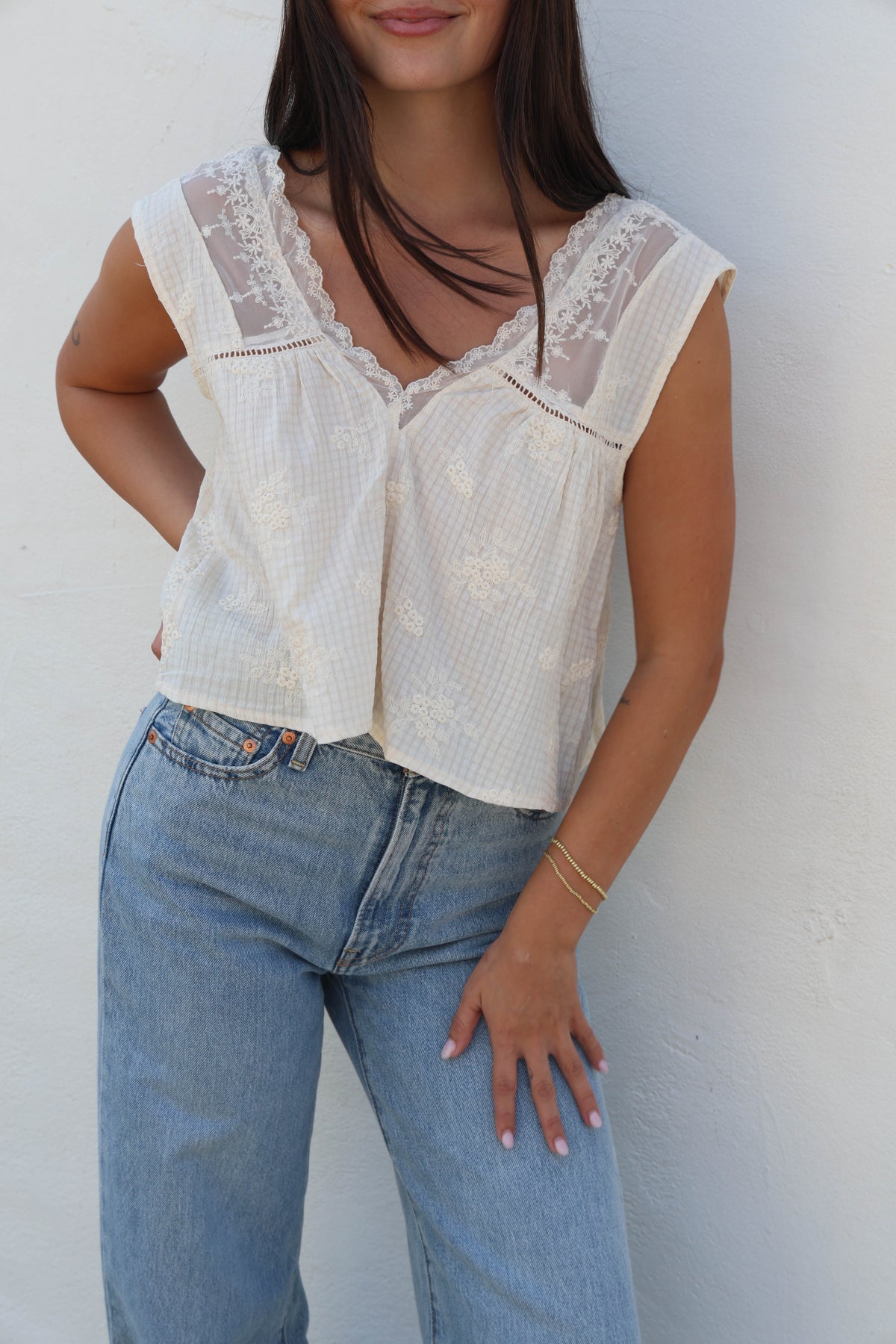 Woman wearing a white tank with lace detail with jeans against a white wall.