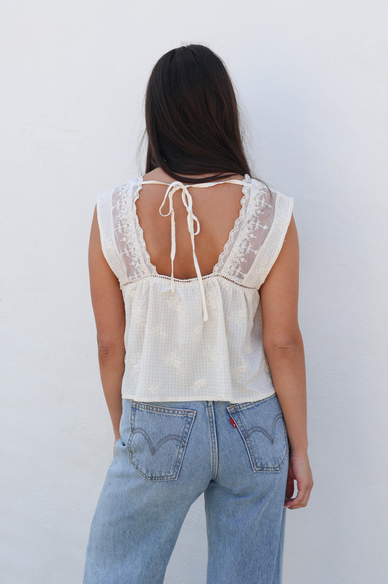 Woman wearing a white tank with lace detail with jeans against a white wall.