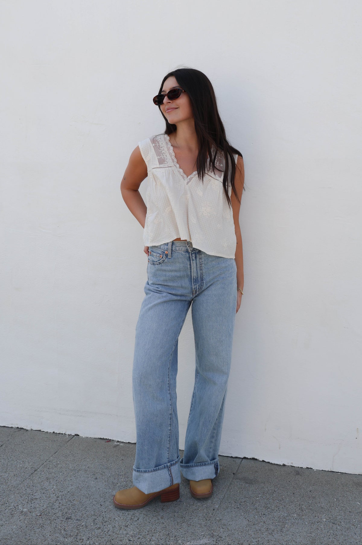 Woman wearing a white tank with lace detail with jeans against a white wall.