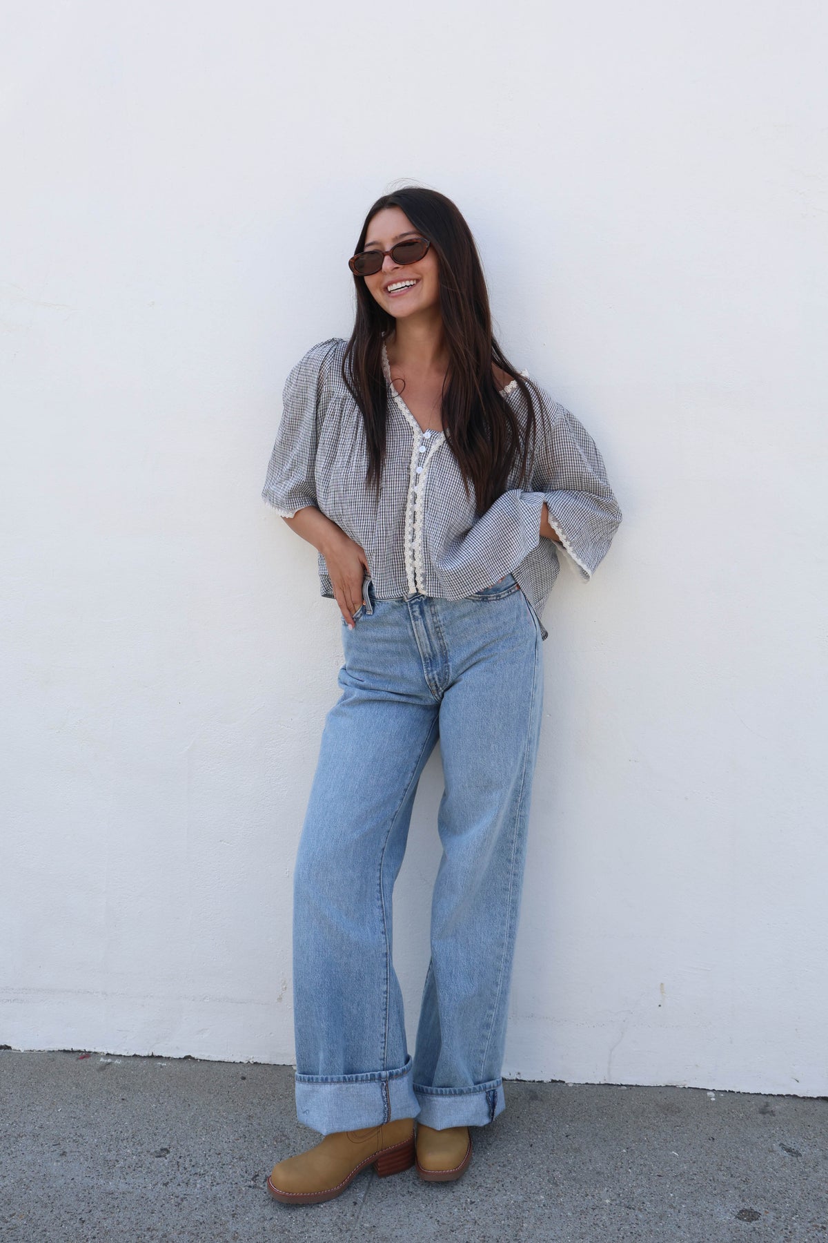 A woman wearing a gray and white gingham puff-sleeve blouse with a white lace-up front, light-wash wide-leg jeans, and tan boots, posing against a plain white wall.