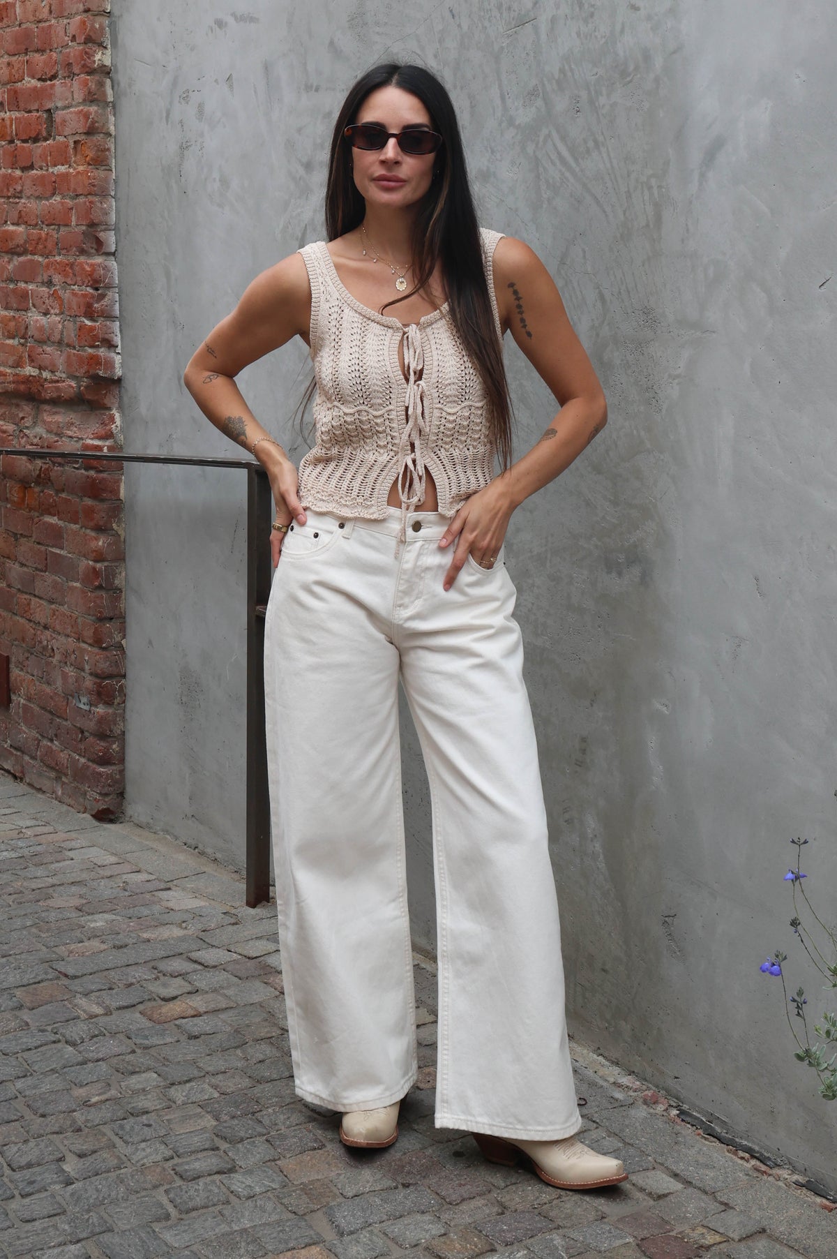 Woman wearing a beige knitted top and white pants standing against a gray wall.