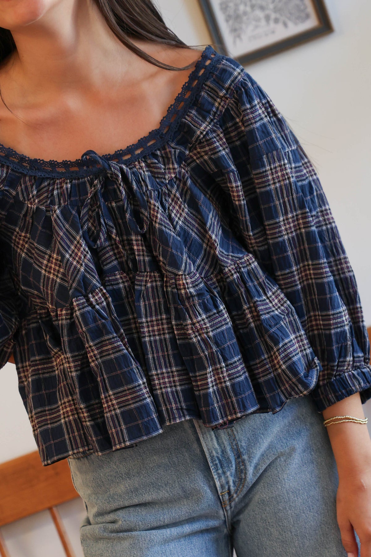 A close-up of a woman wearing a navy blue plaid blouse with a ruffled neckline and tie detail, paired with light blue jeans.