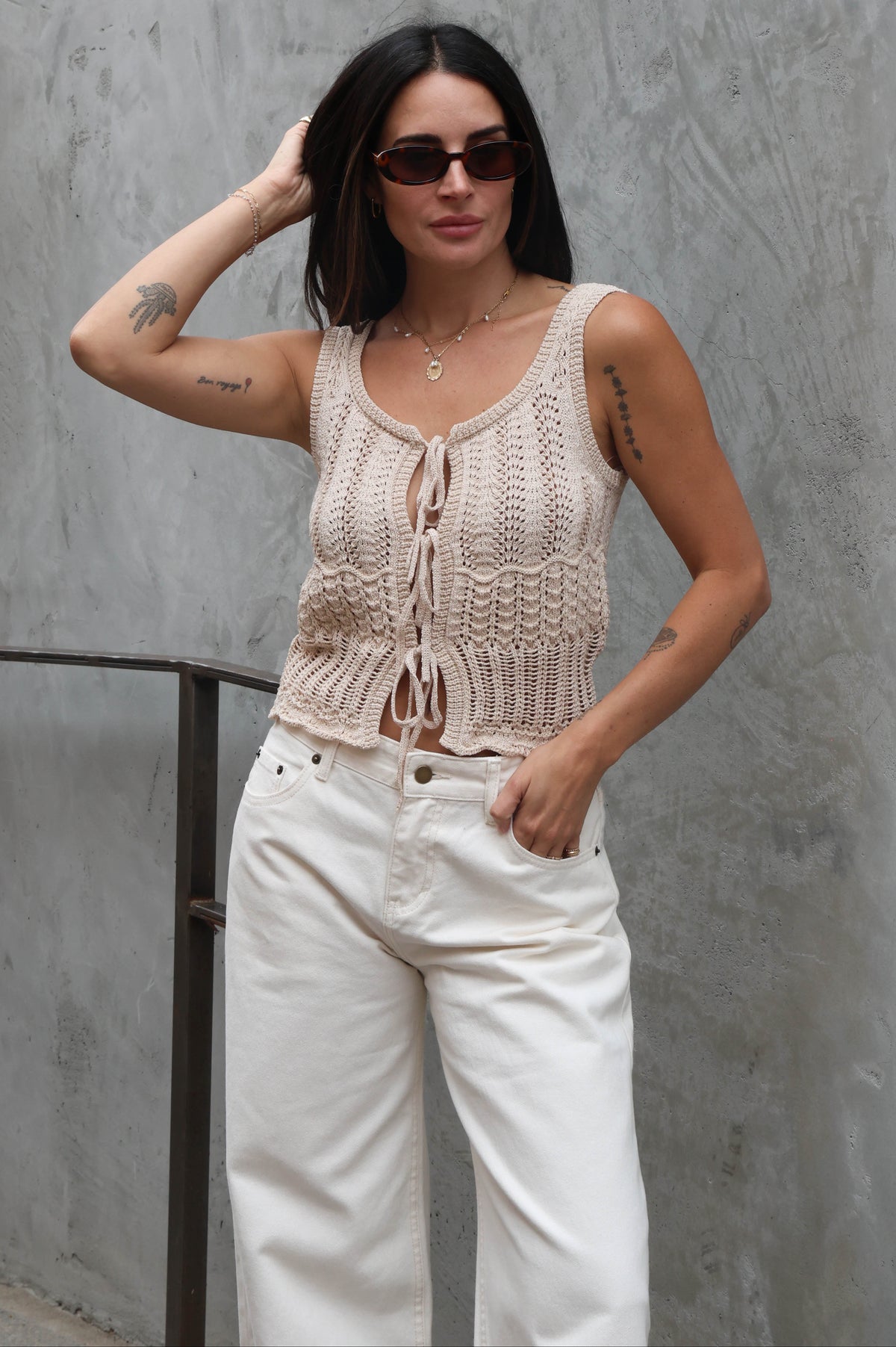 Woman wearing a beige knitted tank top and white pants against a gray wall.
