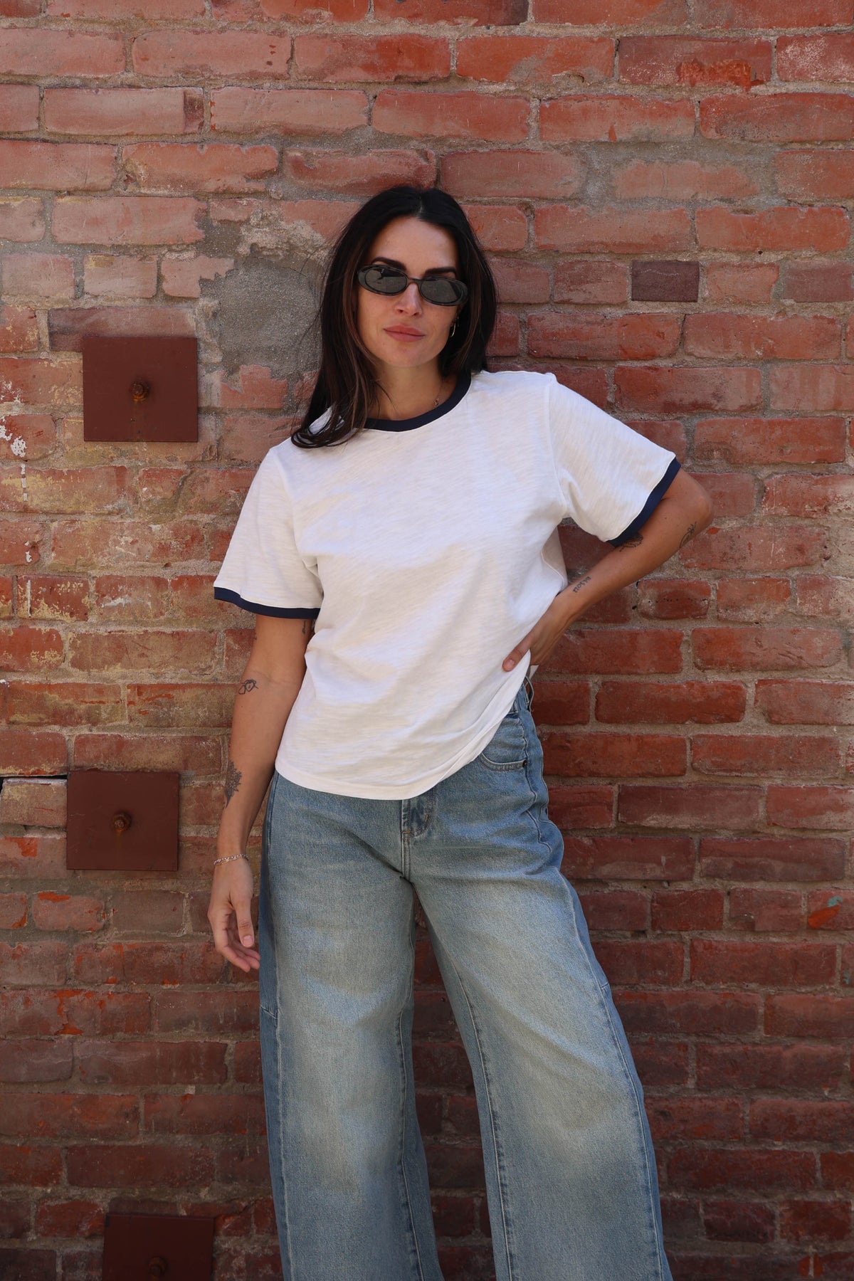 Woman wearing a white t-shirt with jeans against a brick wall.