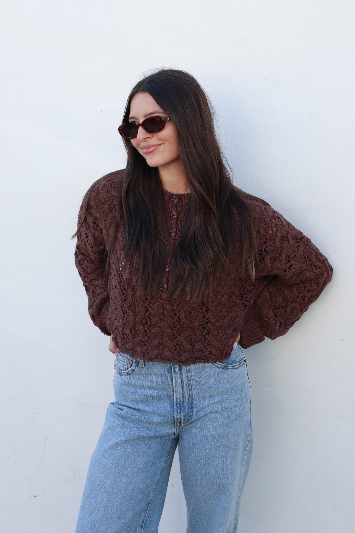 A woman wearing a brown cable-knit sweater, light-wash wide-leg jeans, and tan boots stands against a white wall.