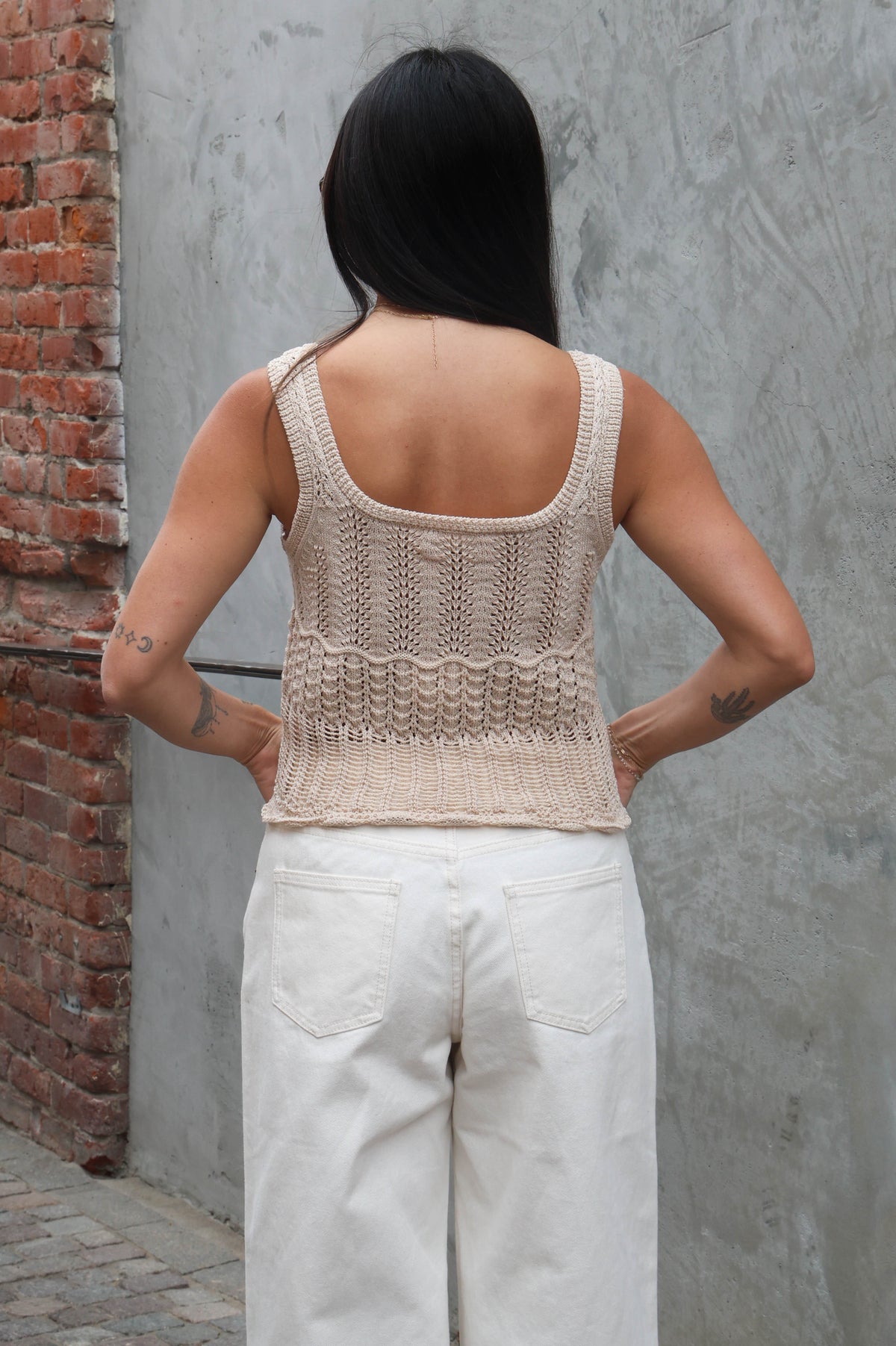 Person wearing a beige knitted tank top and light-colored pants against a gray wall.