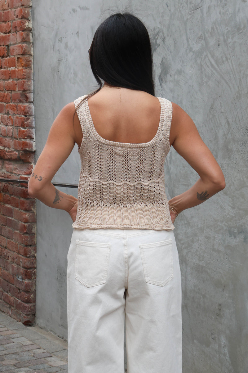 Person wearing a beige knitted tank top and light-colored pants against a gray wall.