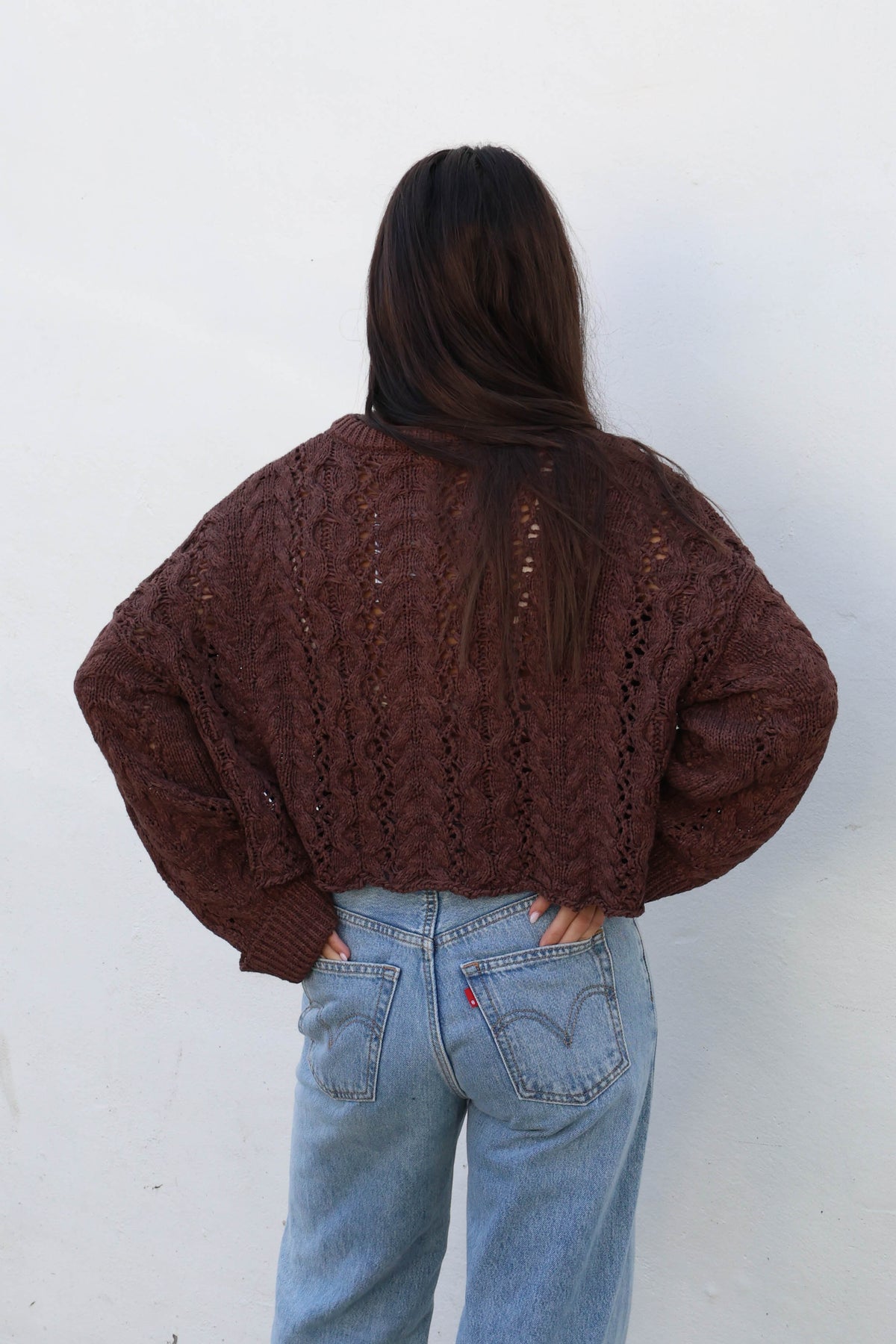 A woman wearing a brown cable-knit sweater, light-wash wide-leg jeans, and tan boots stands against a white wall.