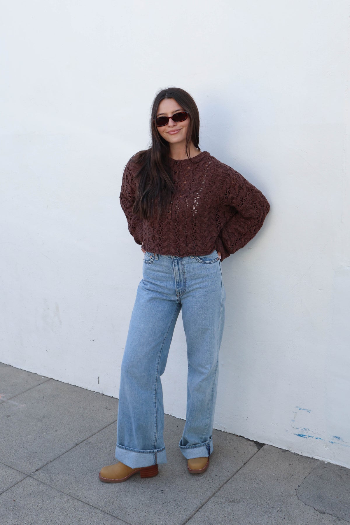 A woman wearing a brown cable-knit sweater, light-wash wide-leg jeans, and tan boots stands against a white wall.