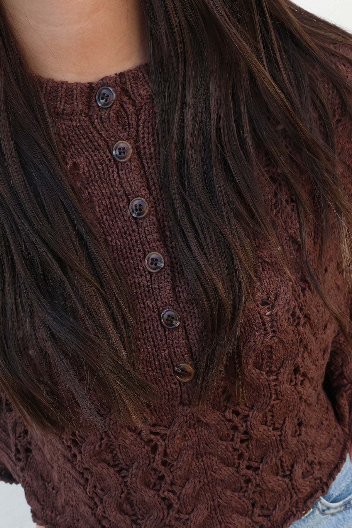 A close-up of a person wearing a brown cable-knit button-front top with long dark brown hair partially covering the front.