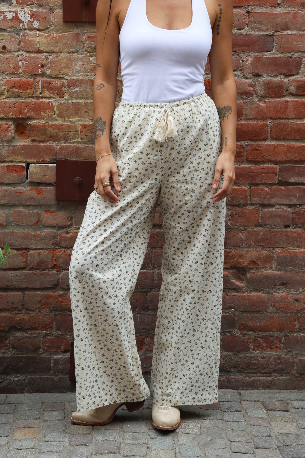 Person wearing a white tank top and patterned pants standing against a brick wall.