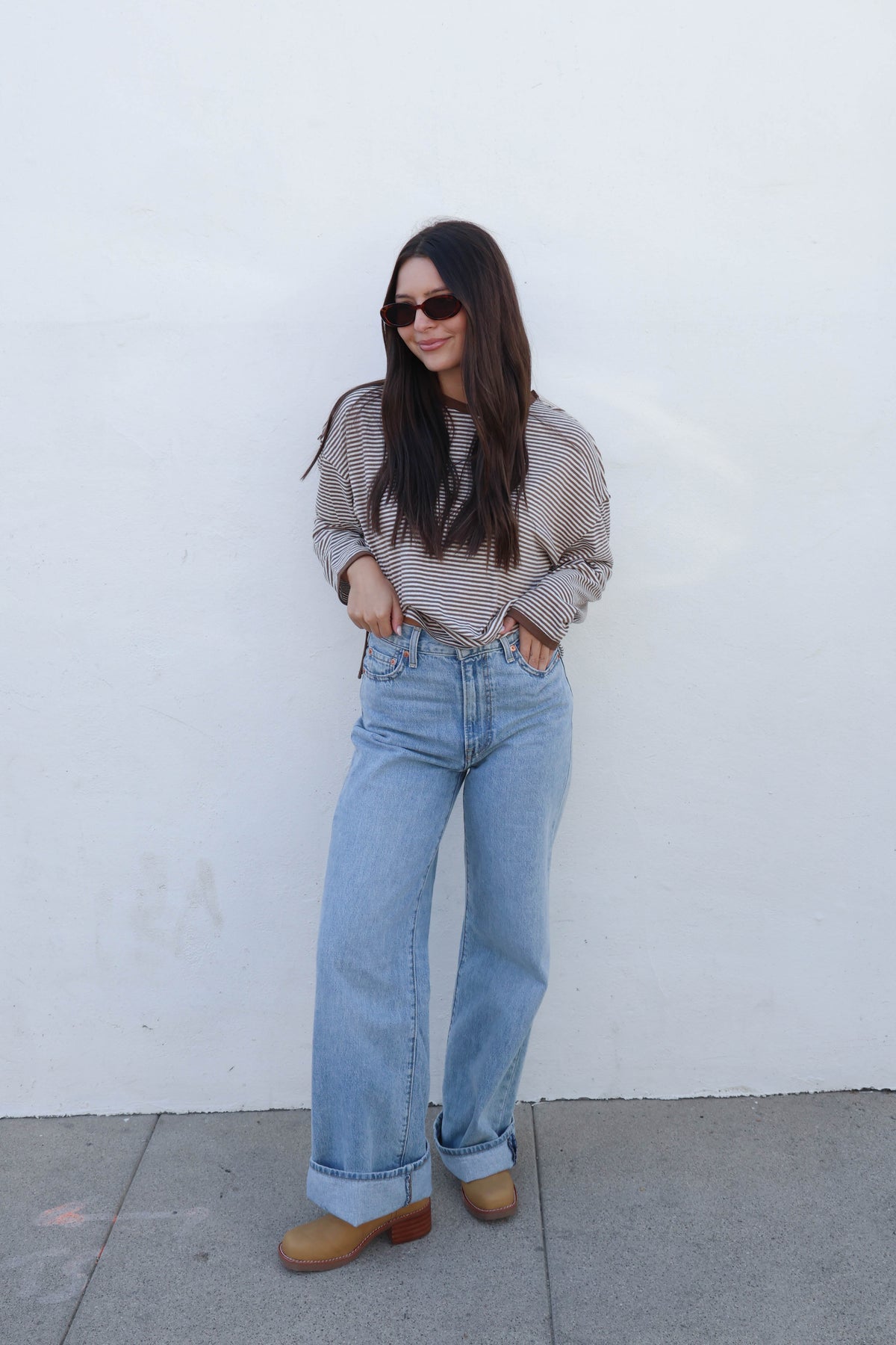 A woman with long dark hair and sunglasses wearing a brown and beige striped long-sleeve crop top and light blue jeans, posing against a white wall.