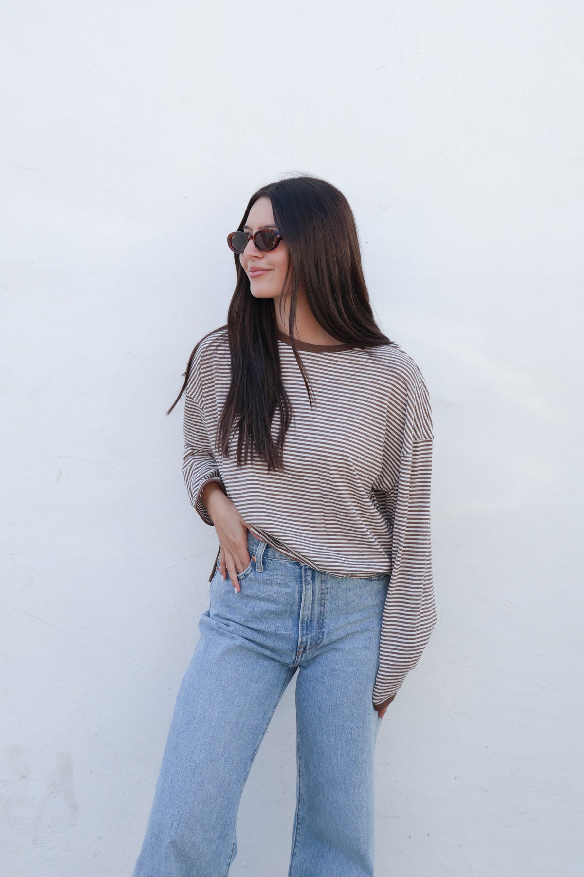 A woman with long dark hair and sunglasses wearing a brown and white striped long-sleeve top and light blue jeans, standing against a plain white wall.