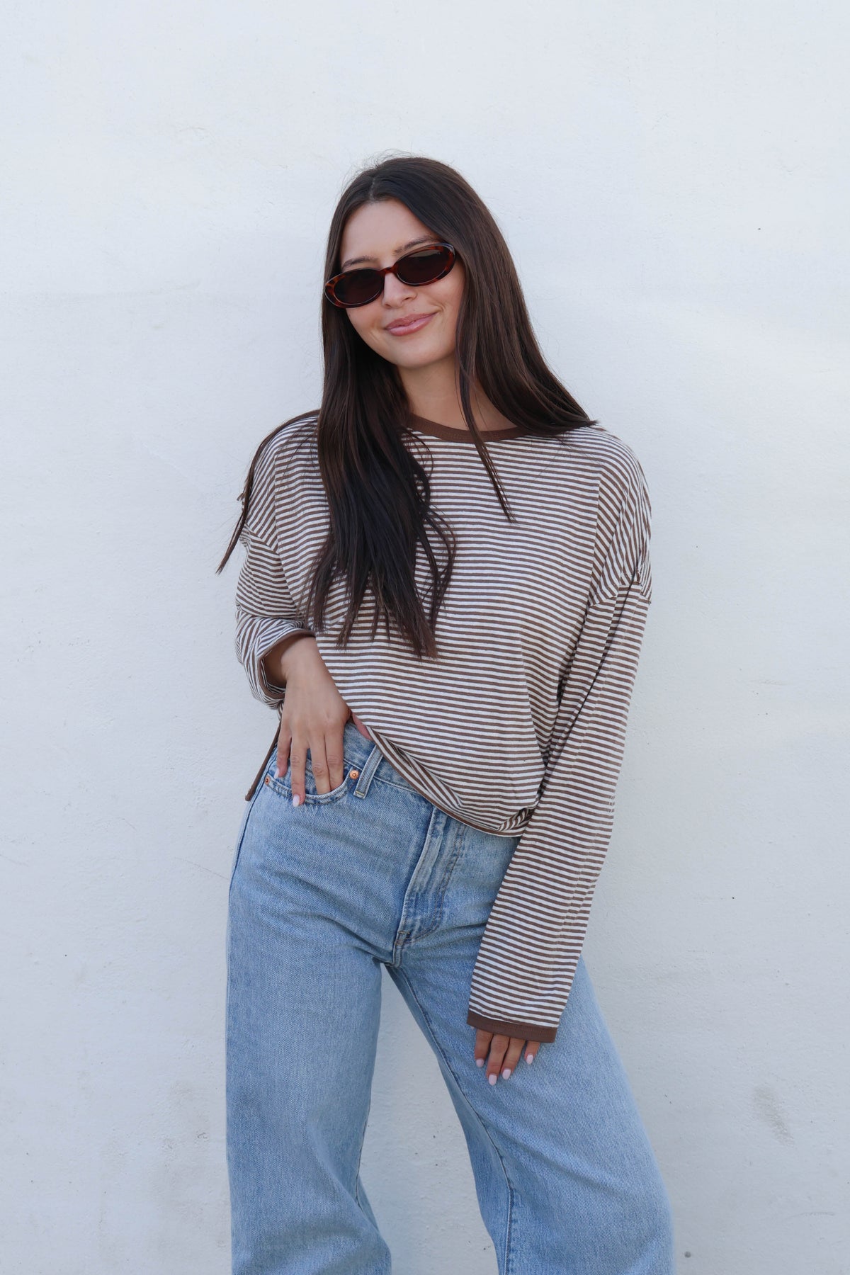A woman with long dark hair and sunglasses wearing a brown and beige striped long-sleeve crop top and light blue jeans, posing against a white wall.