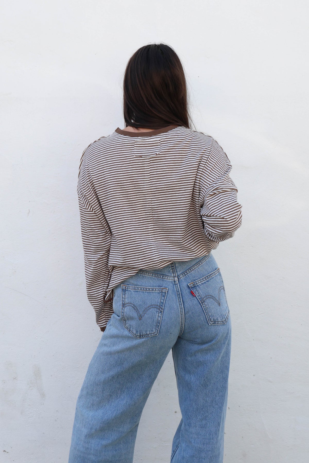 A woman with long dark hair and sunglasses wearing a brown and beige striped long-sleeve crop top and light blue jeans, posing against a white wall.
