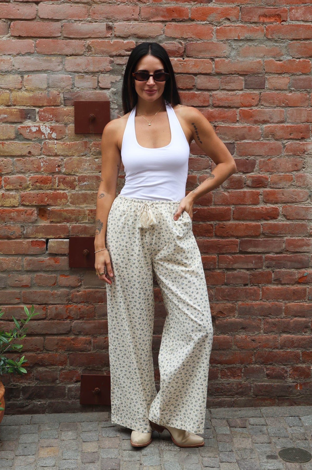 Woman wearing a white top and patterned pants standing against a brick wall.