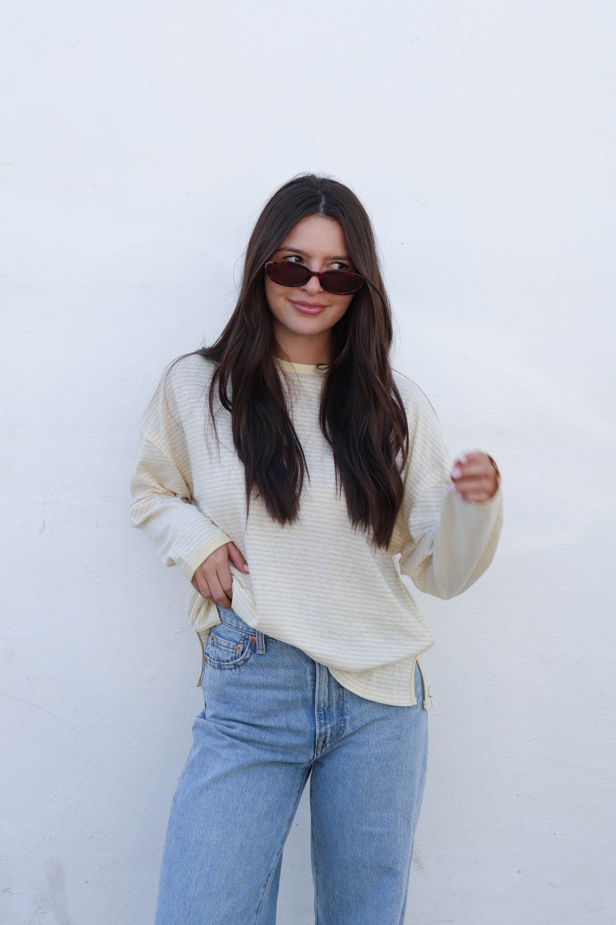 A woman wearing a cream ribbed long-sleeve top and light blue jeans, standing against a plain off-white wall.