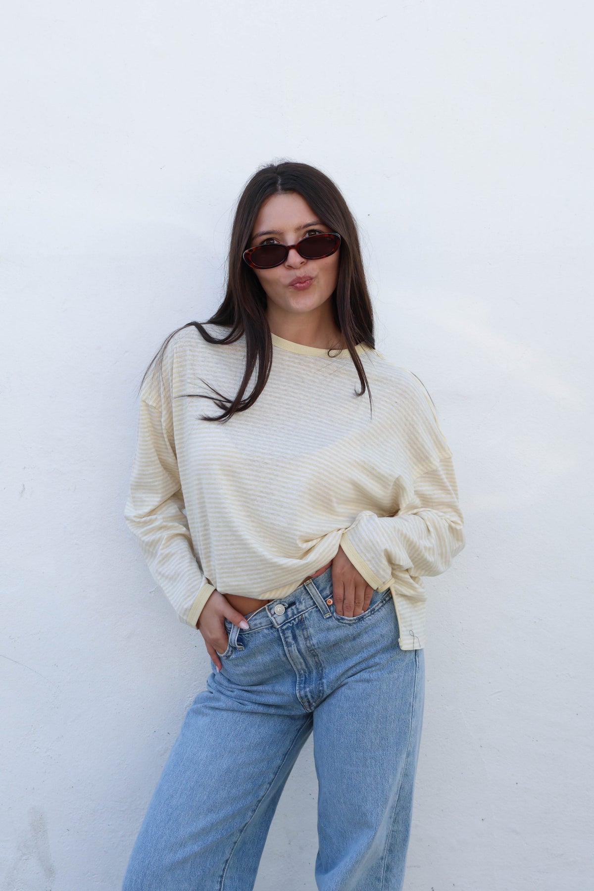 A woman wearing a cream ribbed long-sleeve top and light blue jeans, standing against a plain off-white wall.