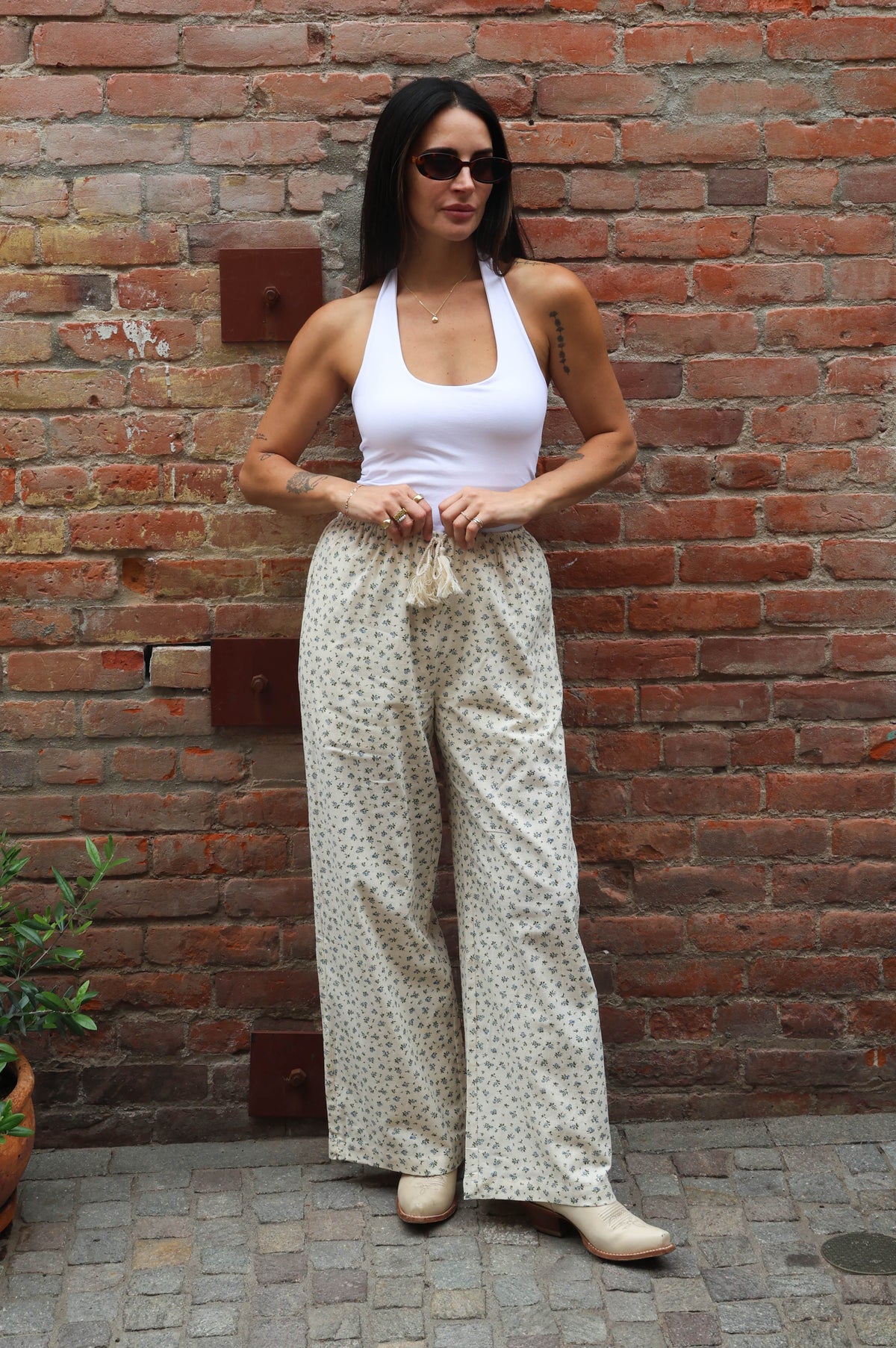 Woman in a white top and patterned pants standing against a brick wall.