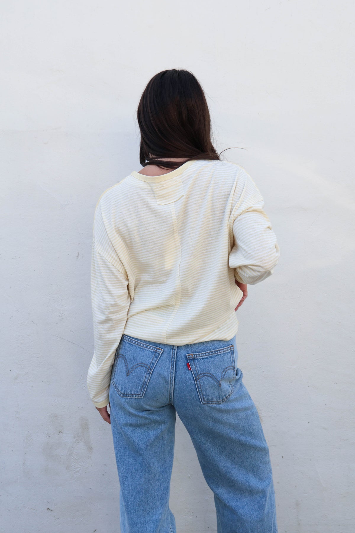 A person shown from behind wearing a cream ribbed long-sleeve top and light blue jeans, standing against a plain off-white wall.