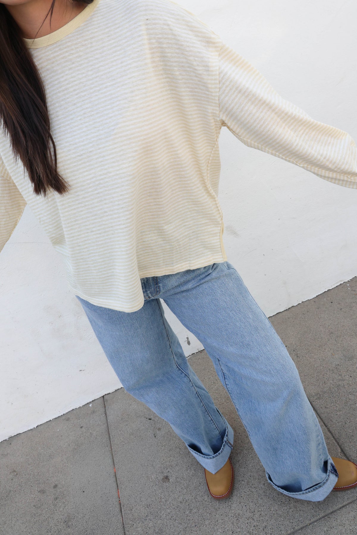 A woman wearing a cream ribbed long-sleeve top and light blue jeans, standing against a plain off-white wall.