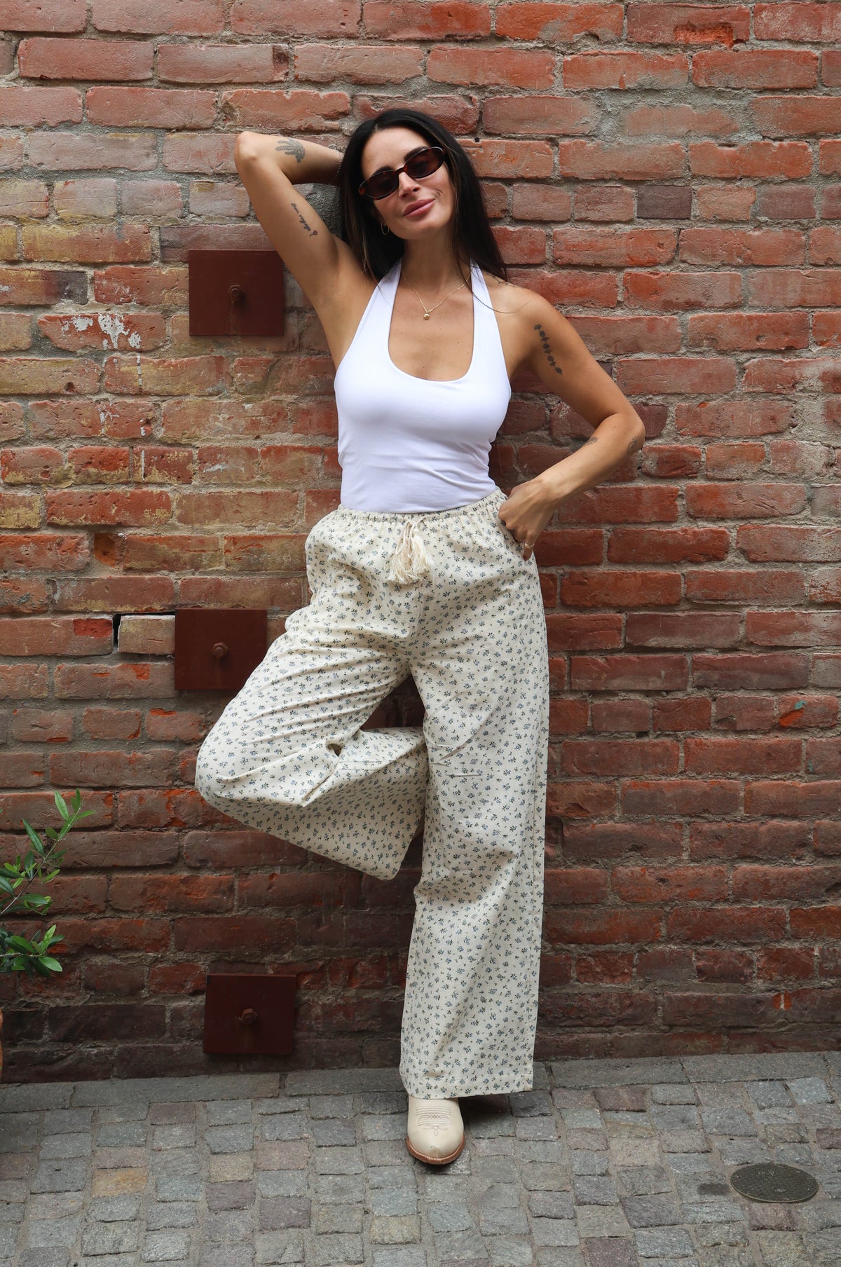 Woman posing against a brick wall wearing a white top and patterned pants.