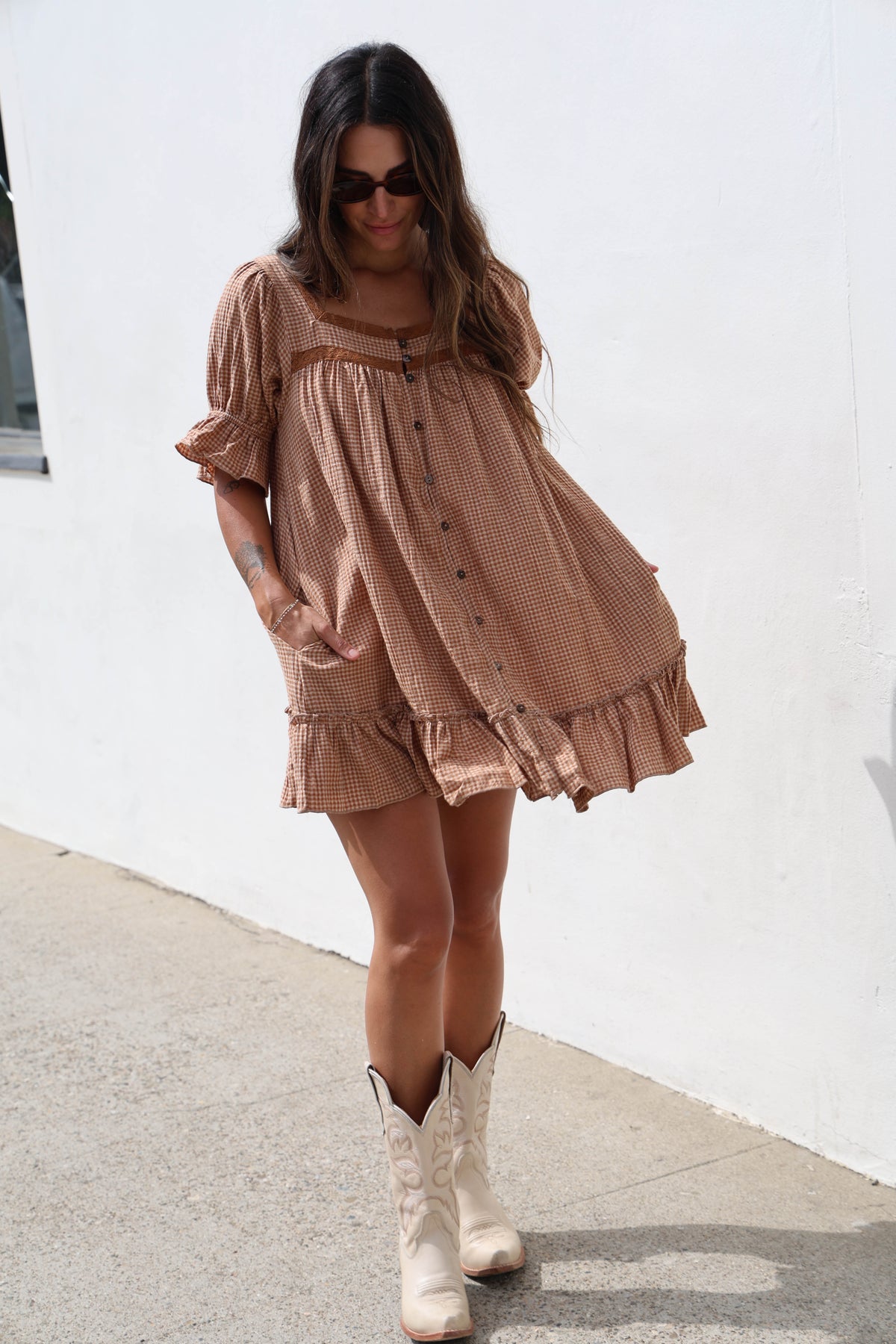 A woman wearing a brown and beige gingham mini dress with puff sleeves and a ruffled hem, paired with cream cowboy boots, standing outdoors against a white wall.