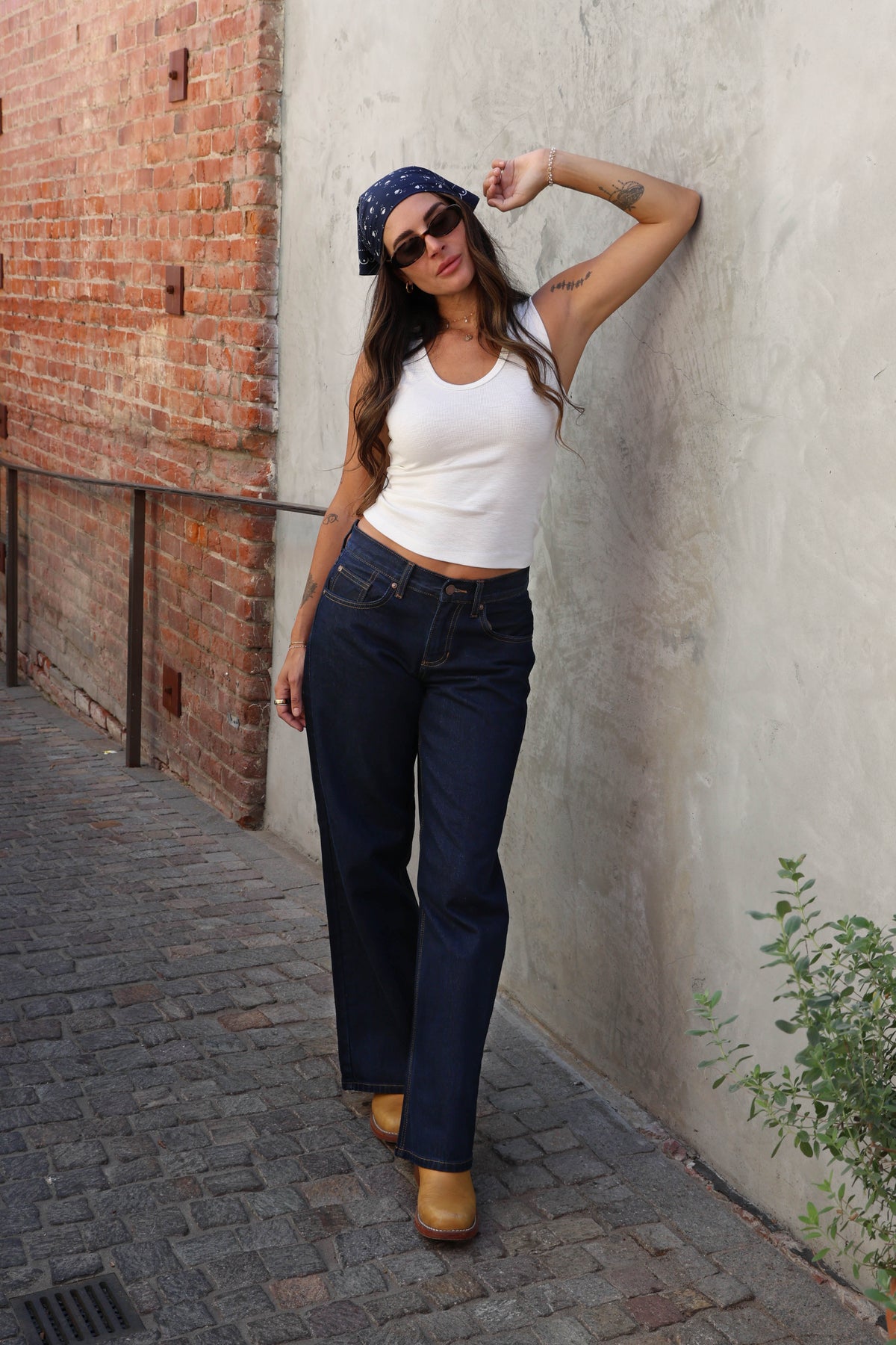 Woman wearing a white top, jeans and a bandana against a plain background.