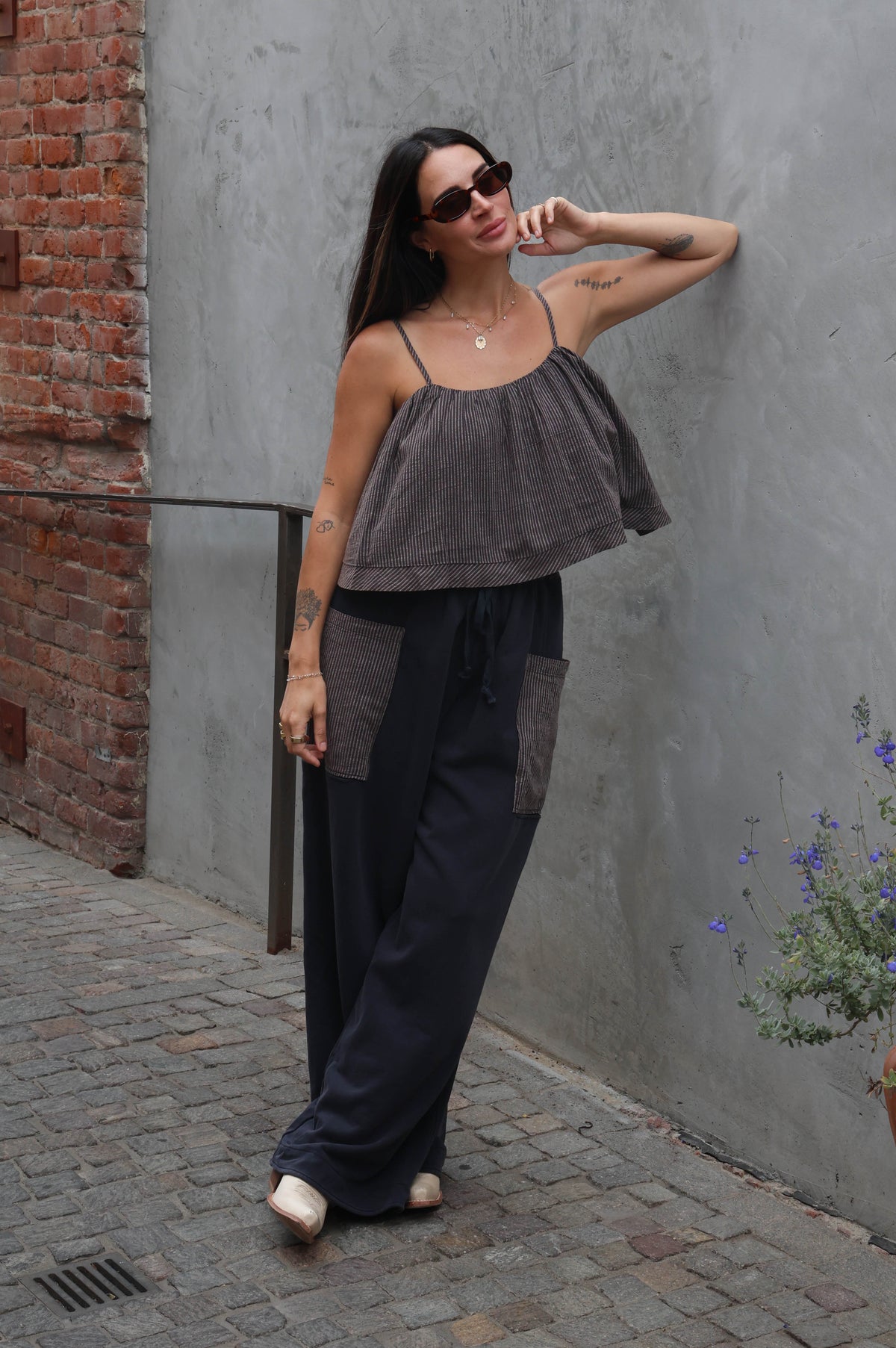 Woman in a gray top and dark pants standing against a gray wall with a brick section.